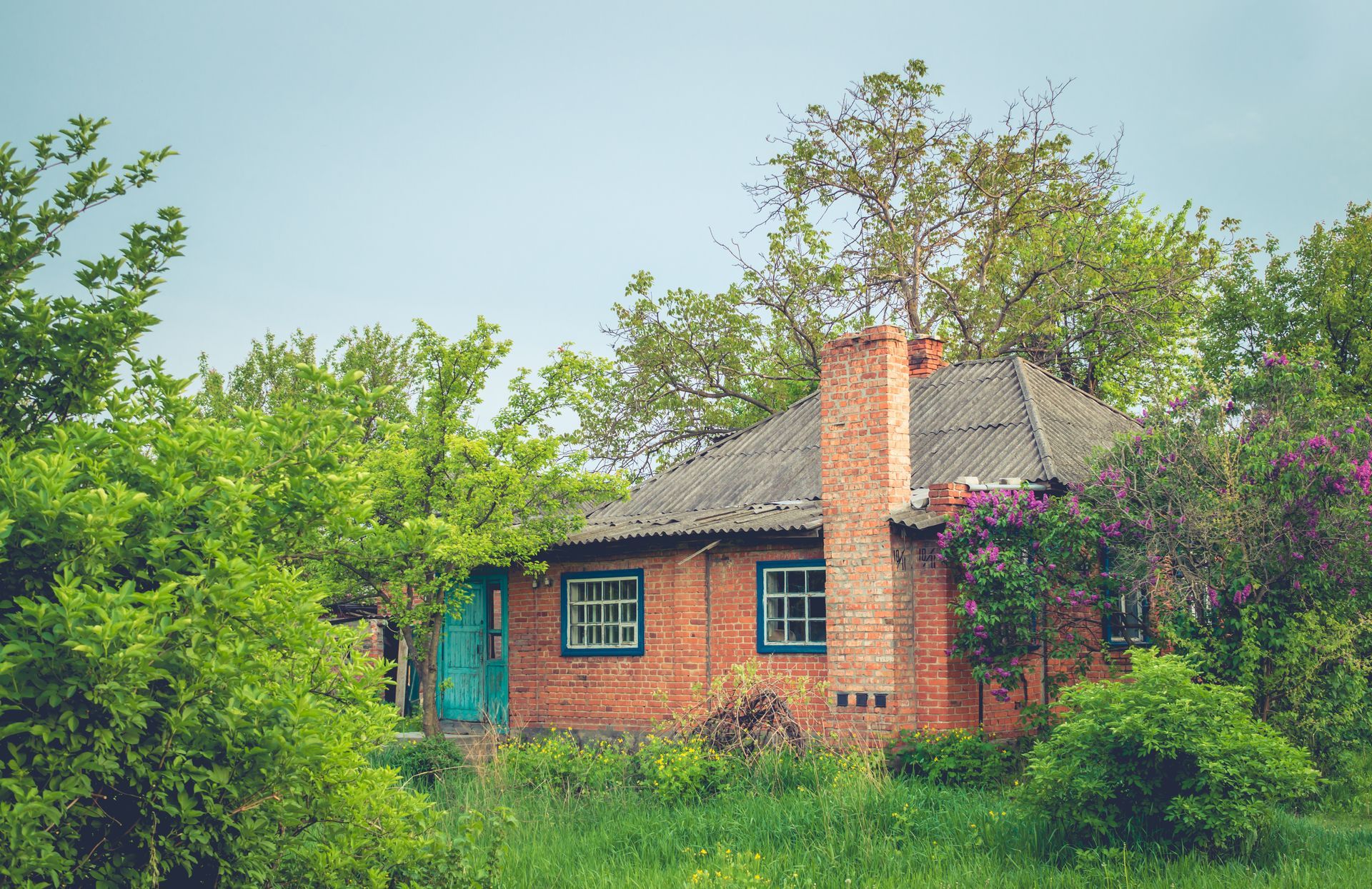 Brick house with chimney, surrounded by green trees and grass, blue windows and door.