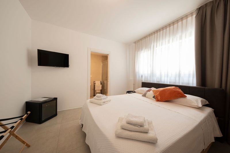 Camera da letto minimalista con letto bianco, testiera nera, tende trasparenti e piccoli comodini.