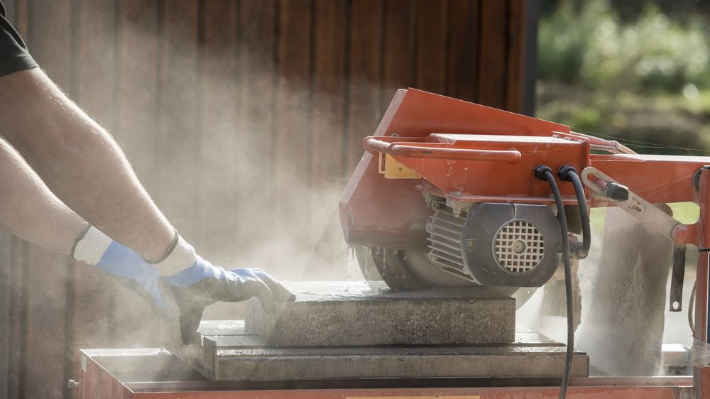 Person Using a Red Saw to Cut a Stone Block, Creating Dust Outdoors — Ultra Services Ground Pavement & Building in Dubbo, NSW