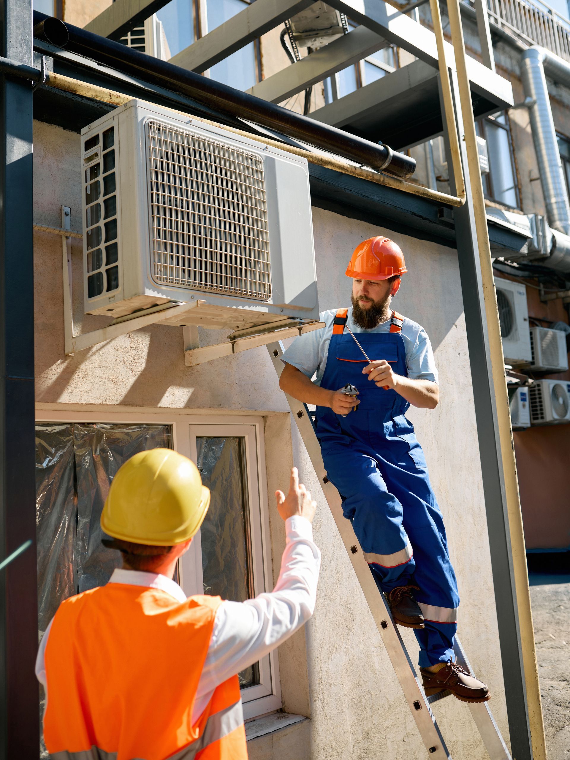 Two workers: One on a ladder, inspecting AC unit; other pointing. Outdoor setting.