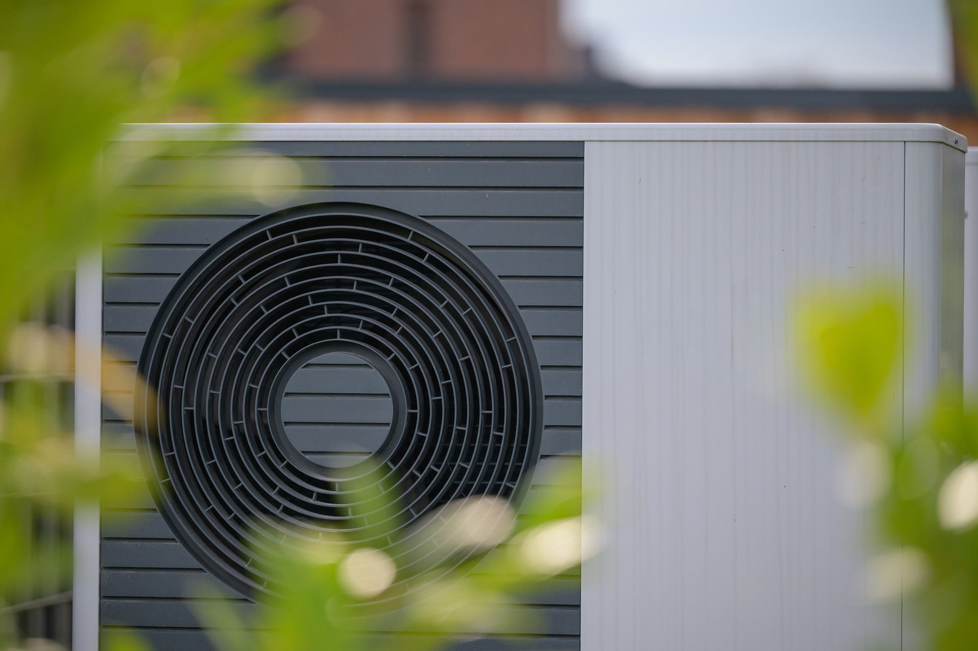 White and dark gray heat pump unit with circular fan, viewed through green foliage.