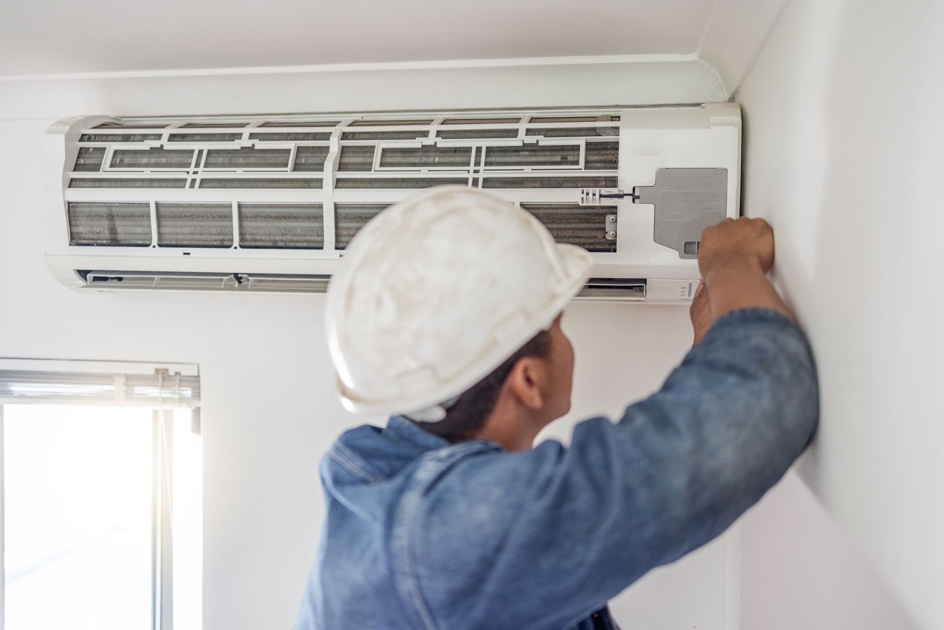 Technician in hard hat servicing an air conditioner on a white wall.