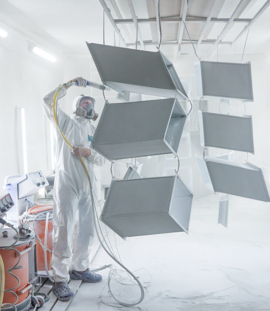 Person in protective suit powder-coating a complex gray metal structure in a white spray booth.