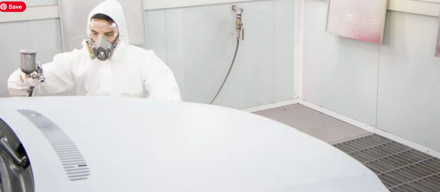 A person wearing a protective suit and mask sprays paint on a car hood inside a paint booth.