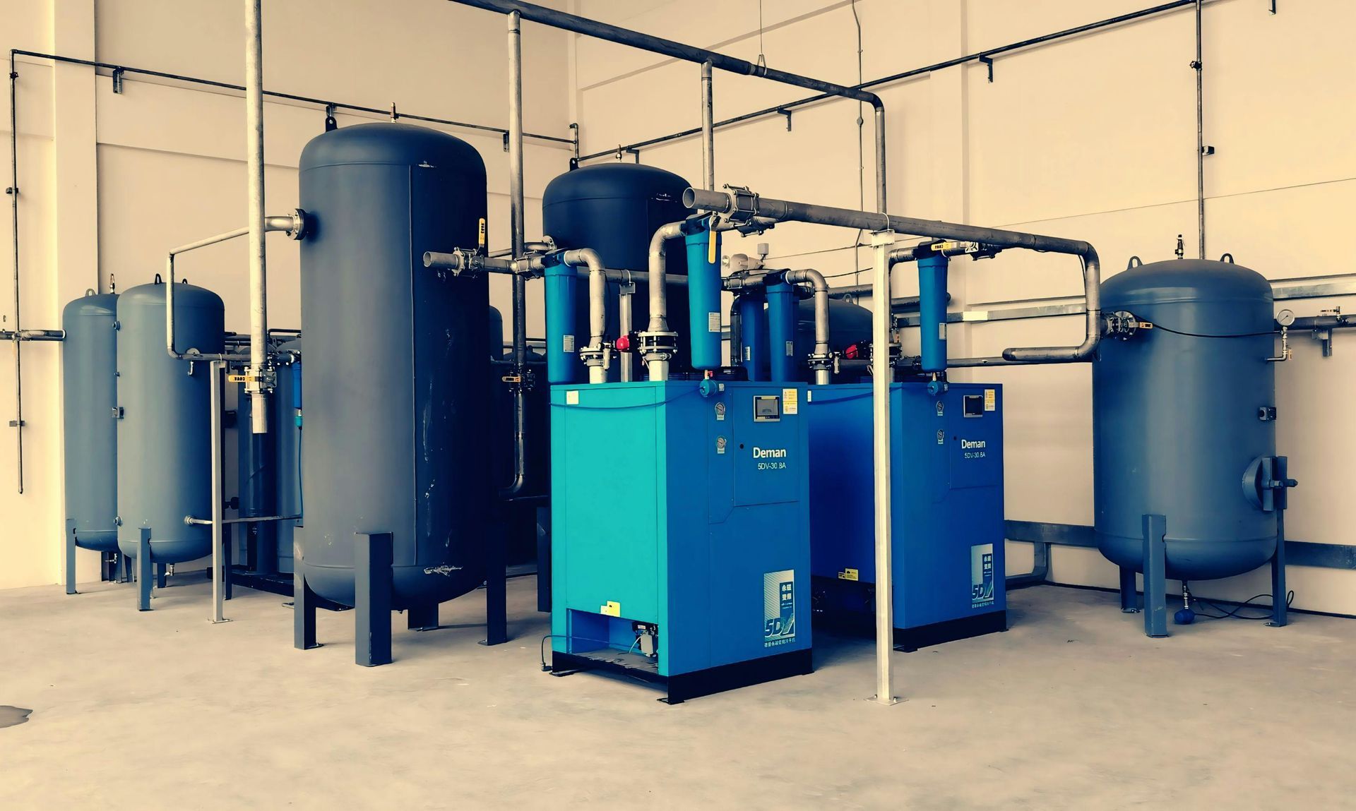 Blue industrial air compressor system in a gray-walled room with various tanks and piping.