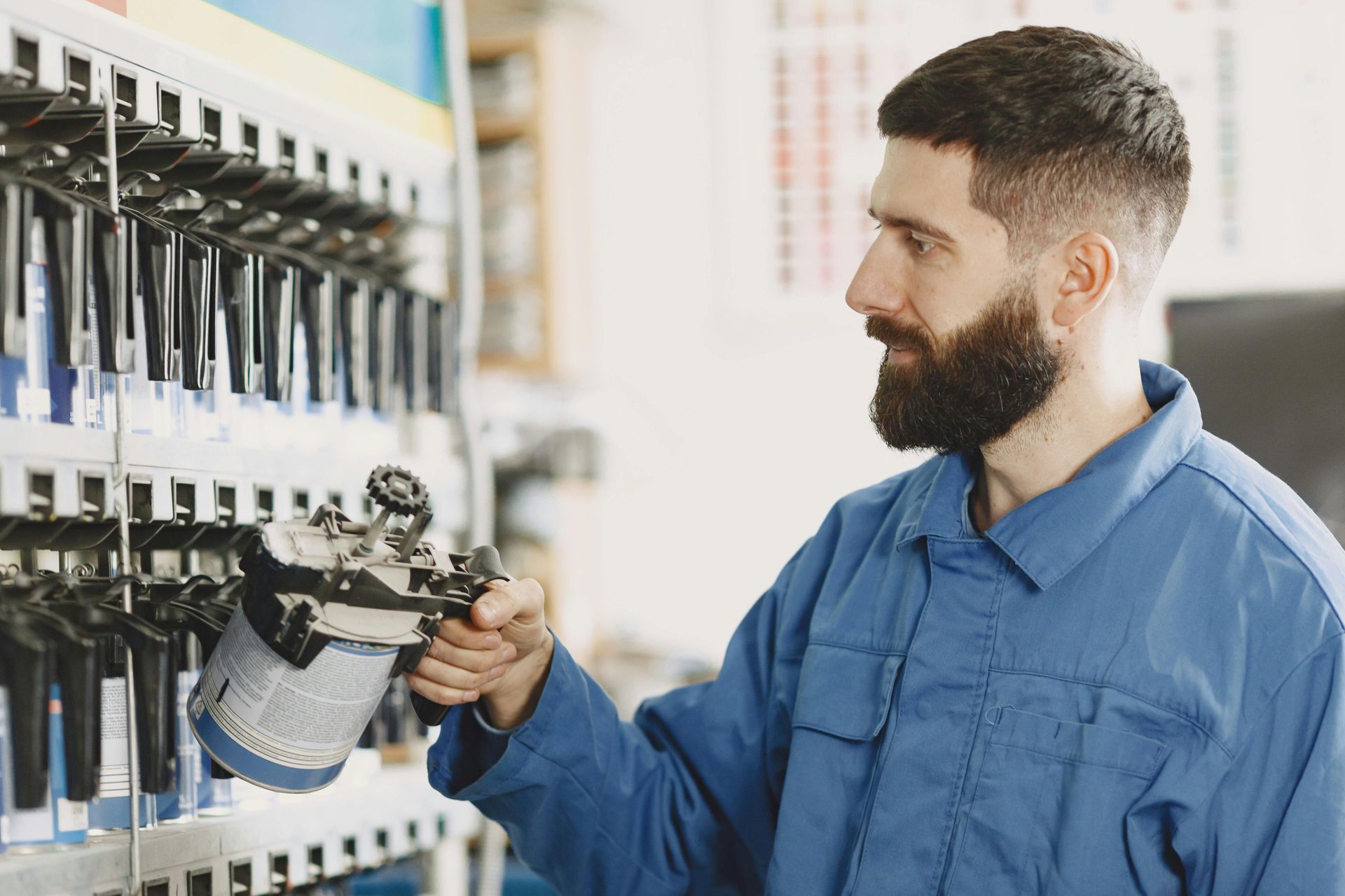 Man in blue jumpsuit holding a spray gun, selecting paint colors in a workshop.