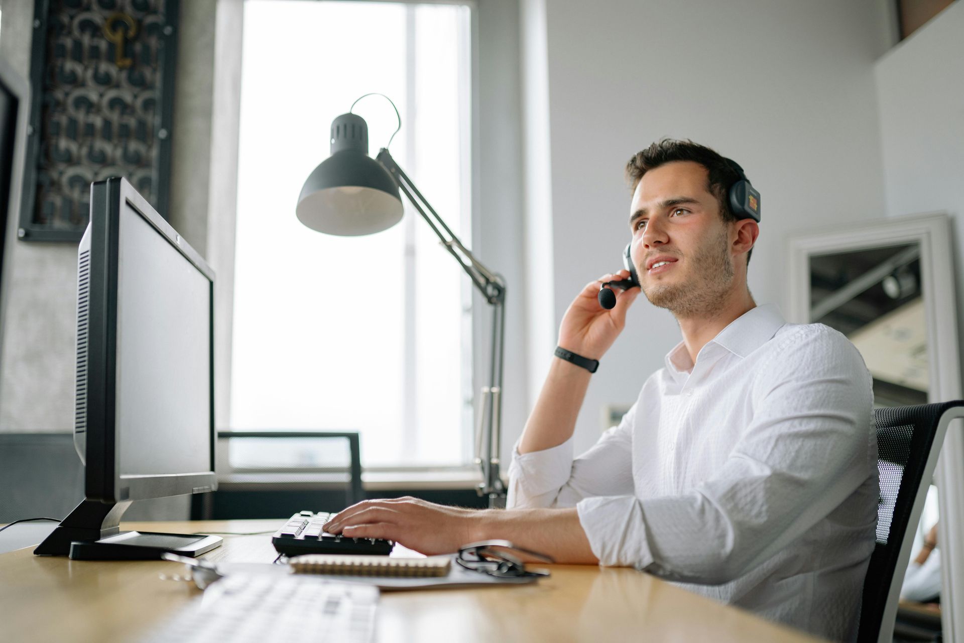 Man wearing a headset, working on a computer at a desk near a window. He appears to be smiling.