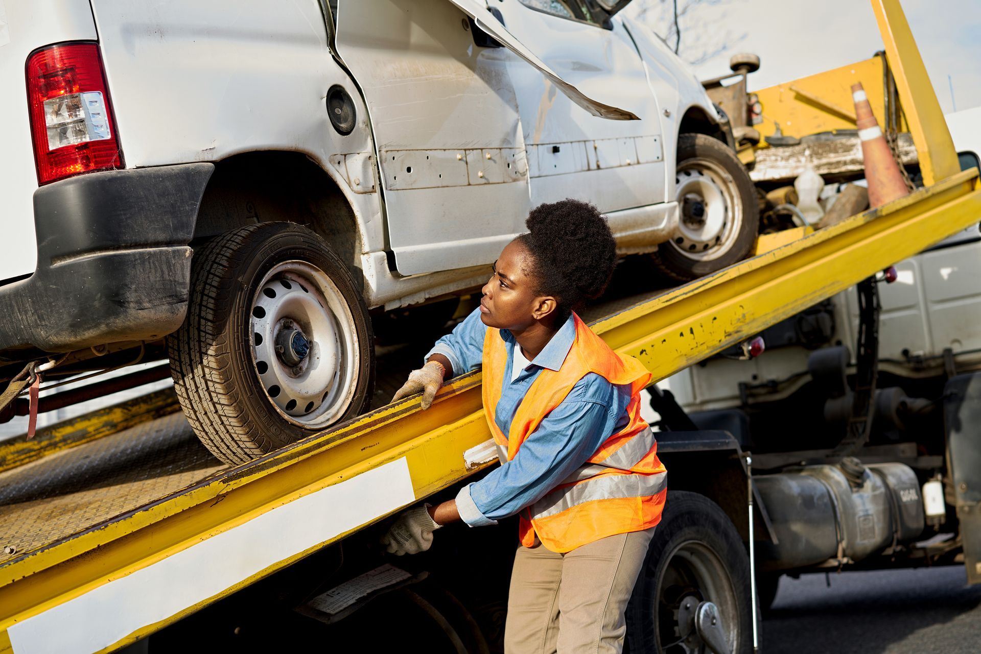 Woman in orange vest guides a damaged white car onto a tow truck.