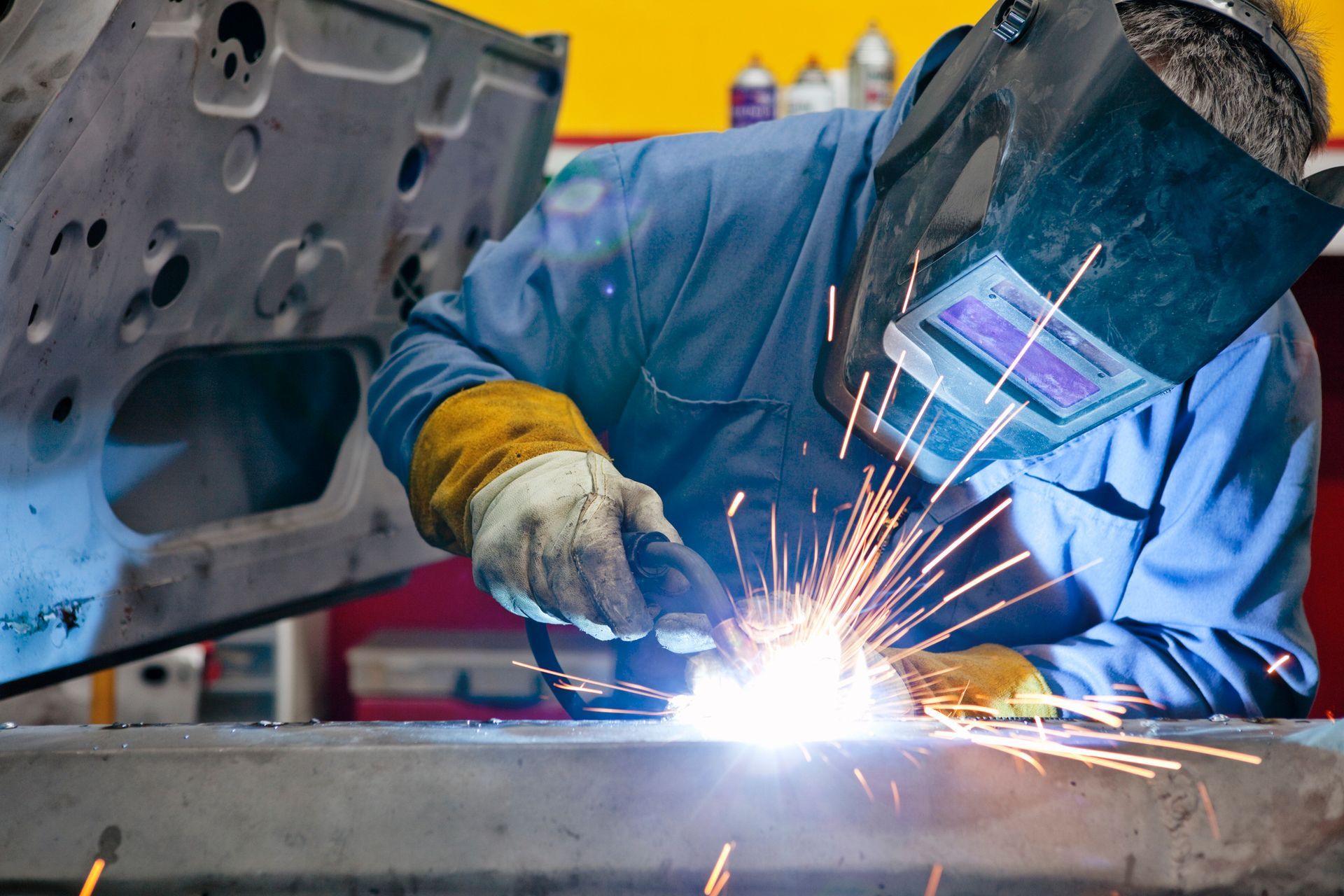Welder in blue coveralls and mask, welding metal with sparks flying.