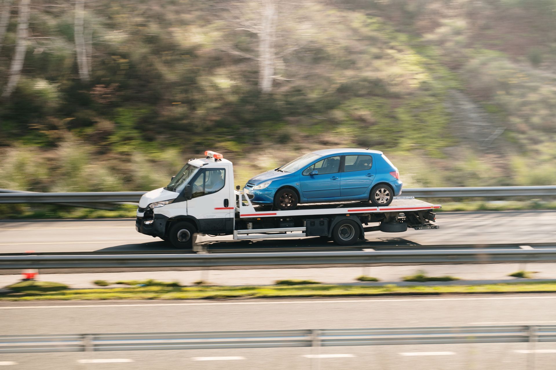 A blue car is being towed on a flatbed truck on a highway, with trees in the background.