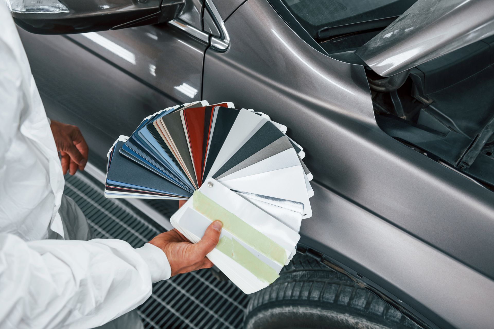 Person in a lab coat holding a color swatch fan next to a gray car, selecting a paint color.