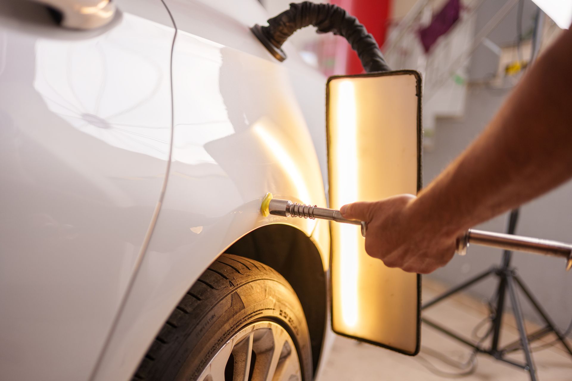 Person using dent removal tool on a white car's panel, lit by a reflector panel, in a garage.