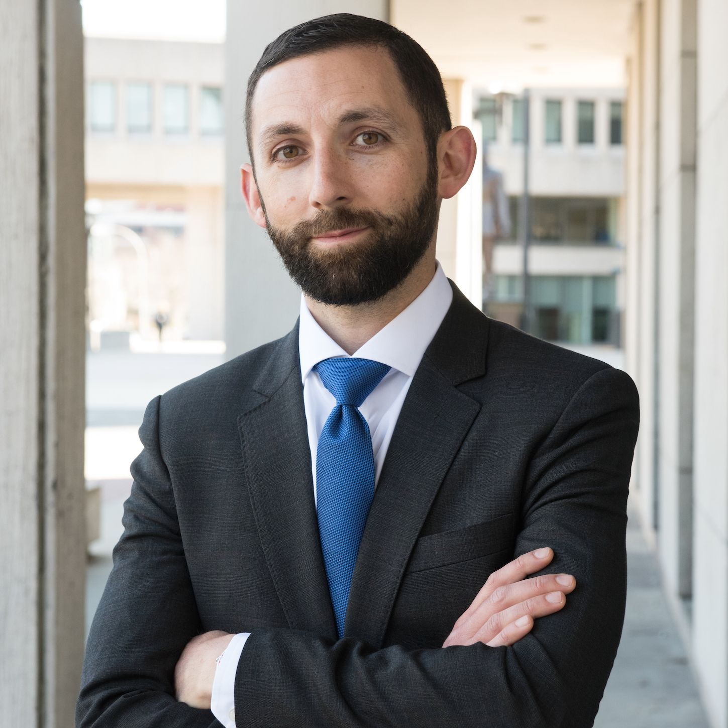 A professional with a short beard, wearing a dark suit and blue tie, stands with arms crossed in an outdoor corridor.