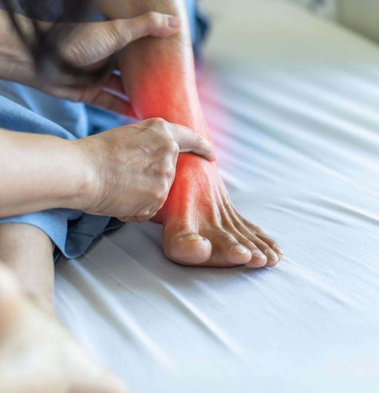 Person touching an ankle, highlighted in red to indicate pain, on a bed.