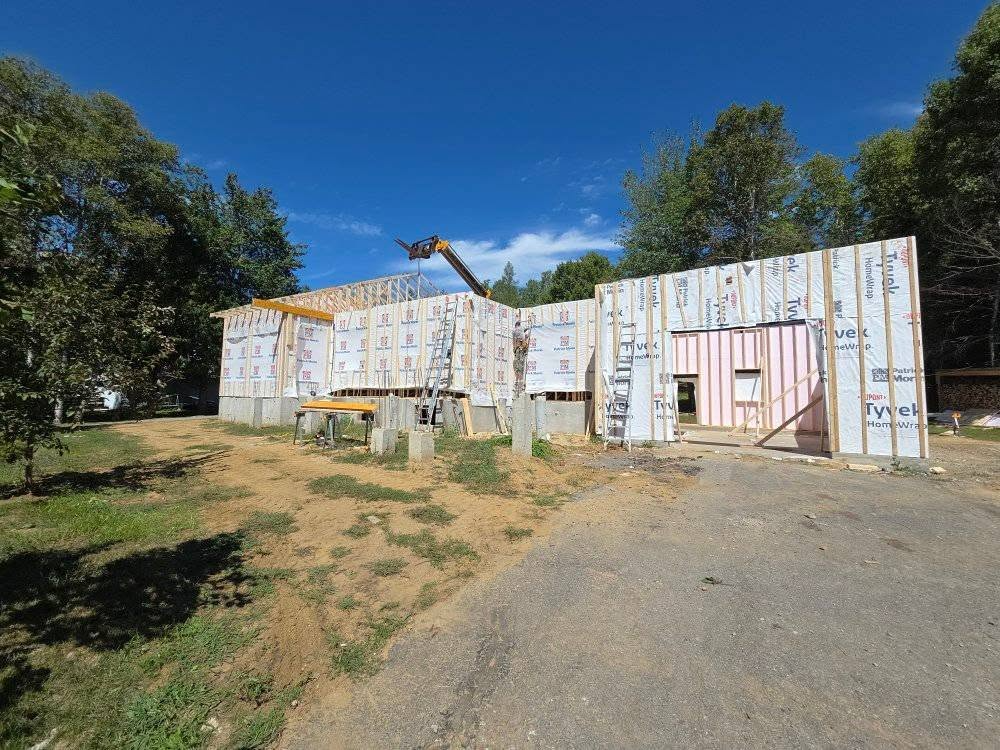 Construction en cours : murs extérieurs à ossature bois, recouverts de revêtement, posés sur des fondations en béton, ciel bleu, arbres en arrière-plan.