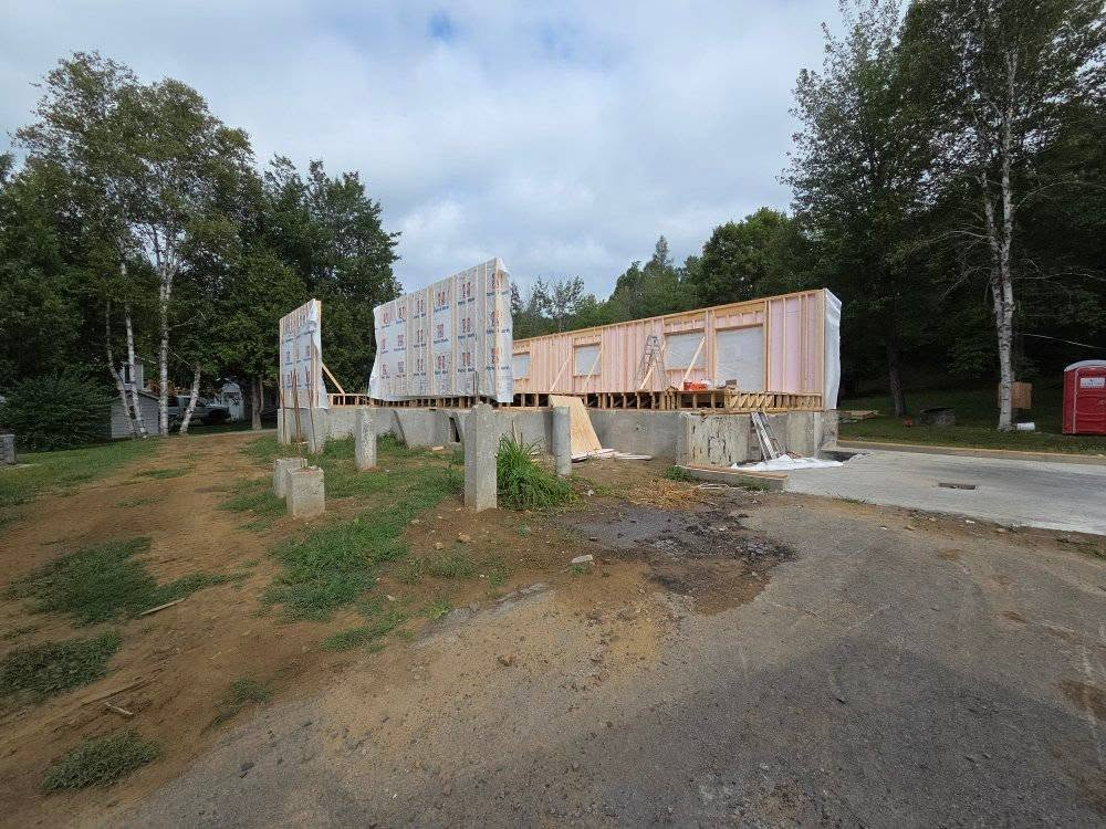 Construction d'une maison sur des fondations en béton, la charpente étant partiellement achevée, entourée d'arbres et de terre.