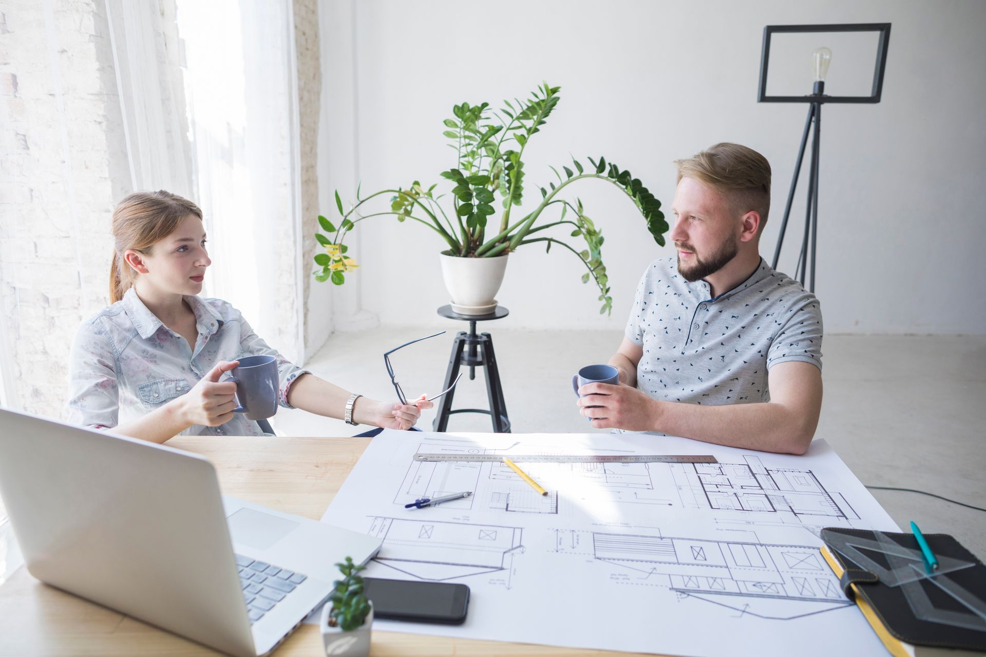 Un homme et une femme assis à une table, discutant de plans, tenant des tasses, avec un ordinateur portable et une plante dans une pièce lumineuse.