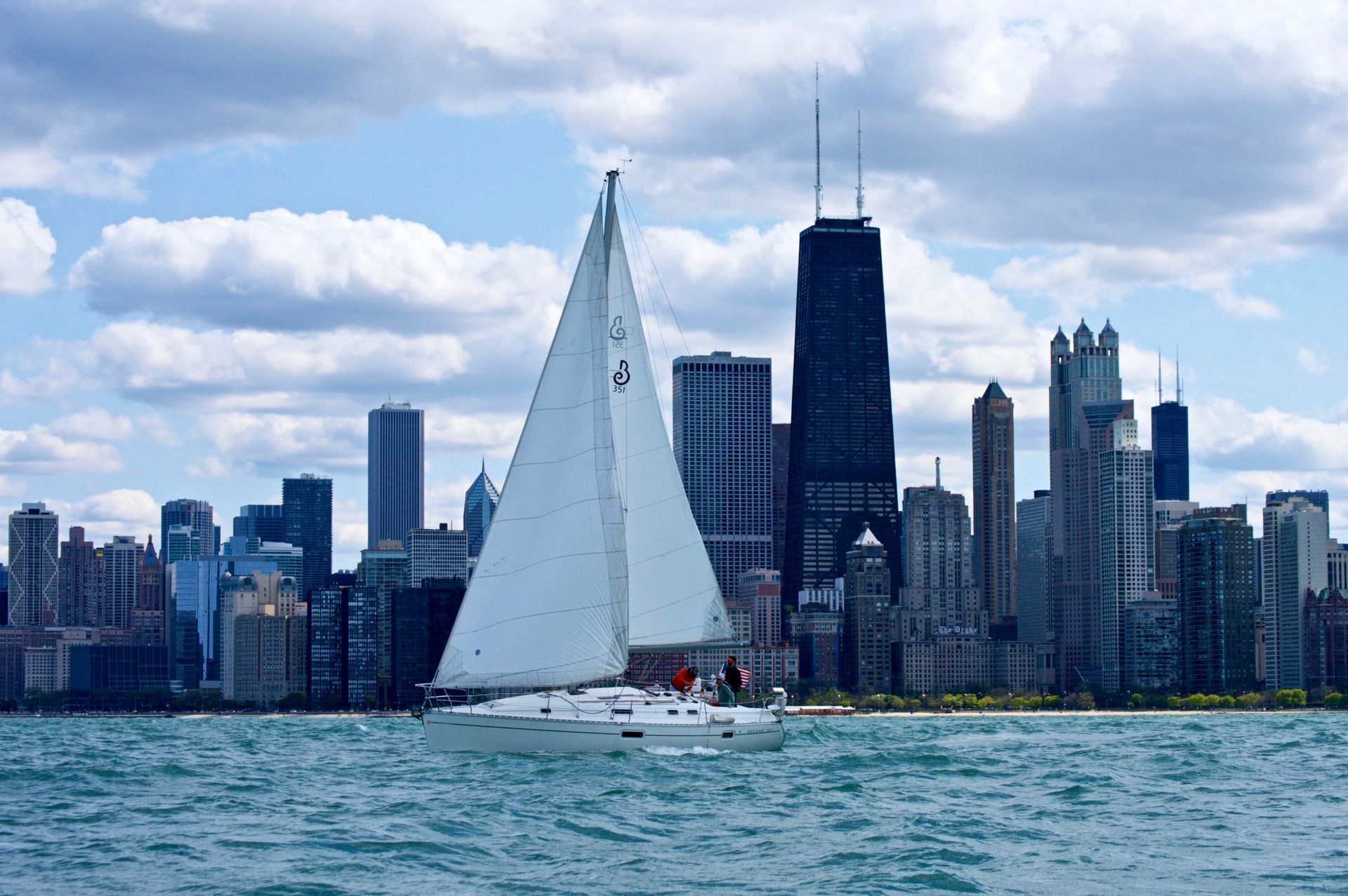 A sailboat with white sails crosses a choppy, turquoise lake in front of the Chicago skyline featuring the Hancock Center.