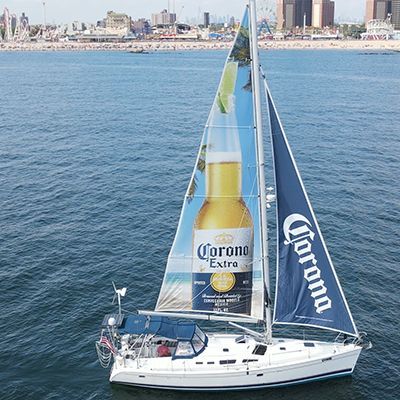 Sailboat with Corona Extra branding on sails, blue water, beach in the background.