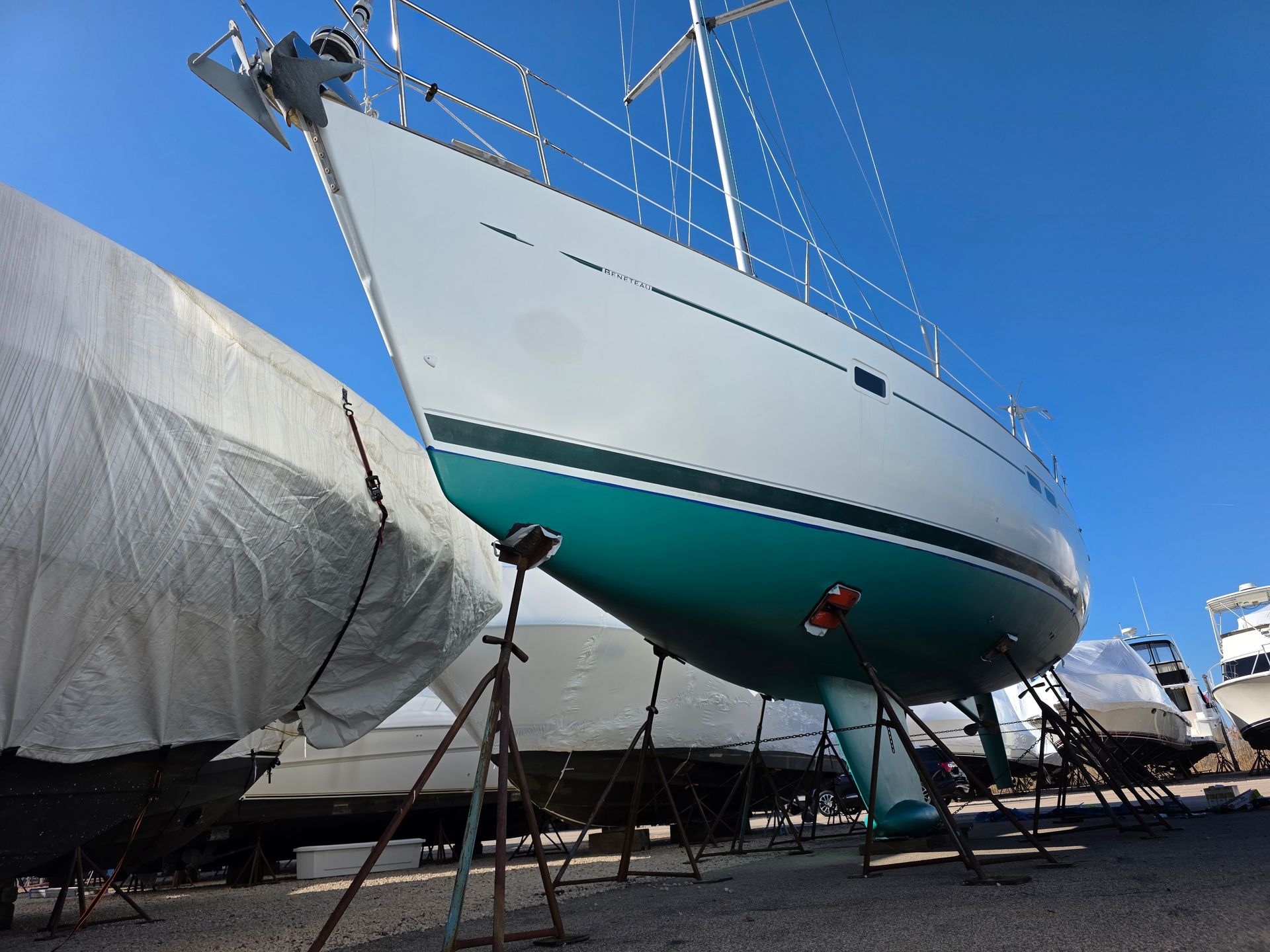 A sailboat hull with a white upper section and teal bottom paint, supported by metal stands in a boat storage yard.