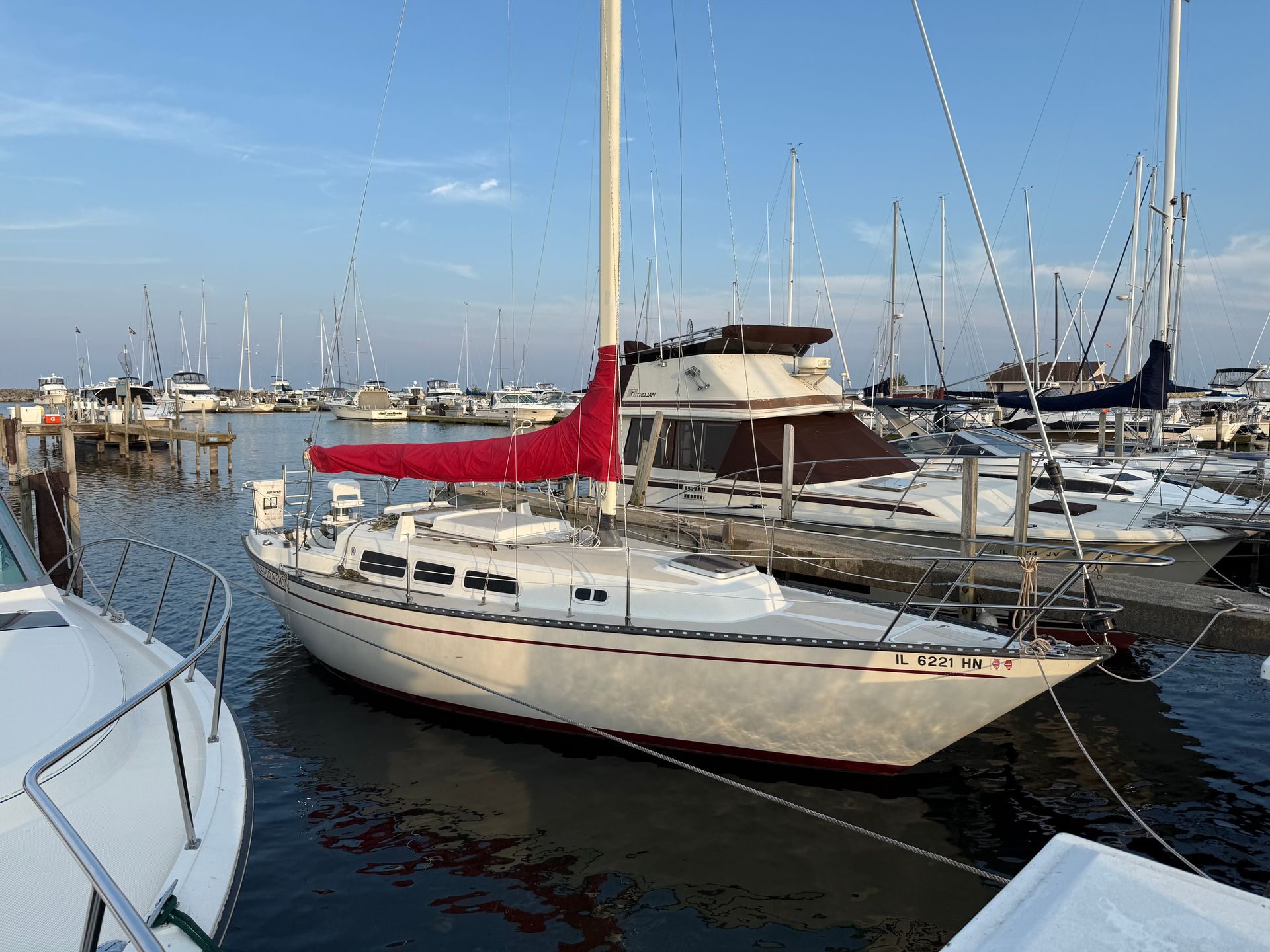 A white sailboat with a red mainsail cover docked at a marina during the day, surrounded by other boats and docks.