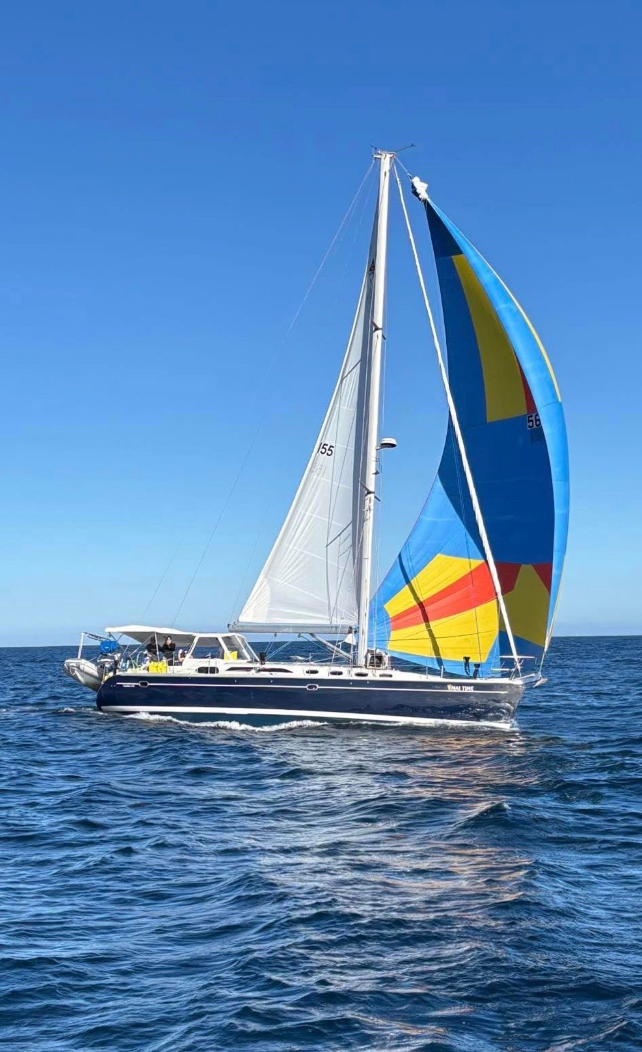 A sailboat with a colorful blue, yellow, and orange spinnaker sailing on blue water under a clear sky.