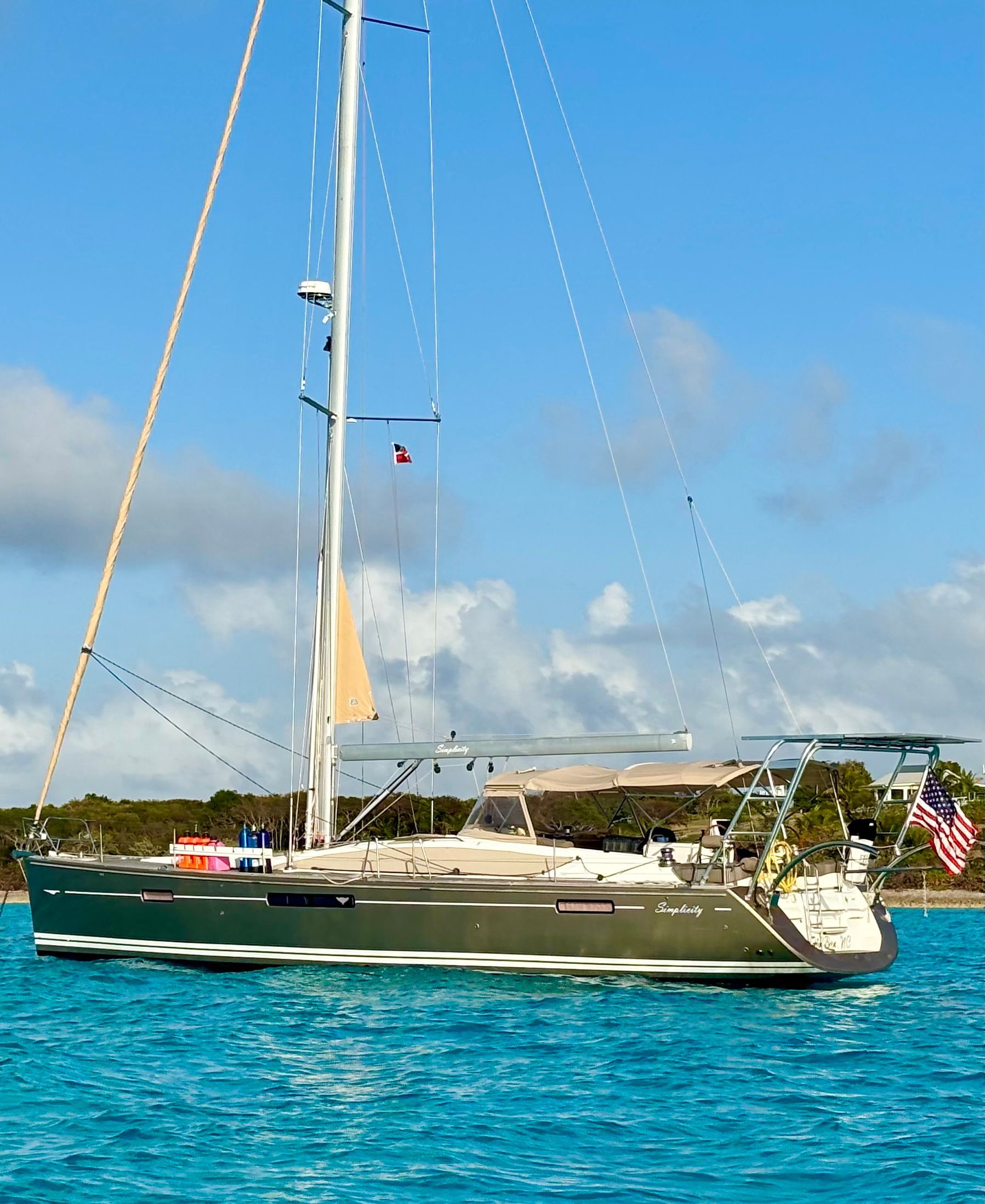 A dark-hulled sailboat with a mast and an American flag docked in calm, bright blue tropical water.