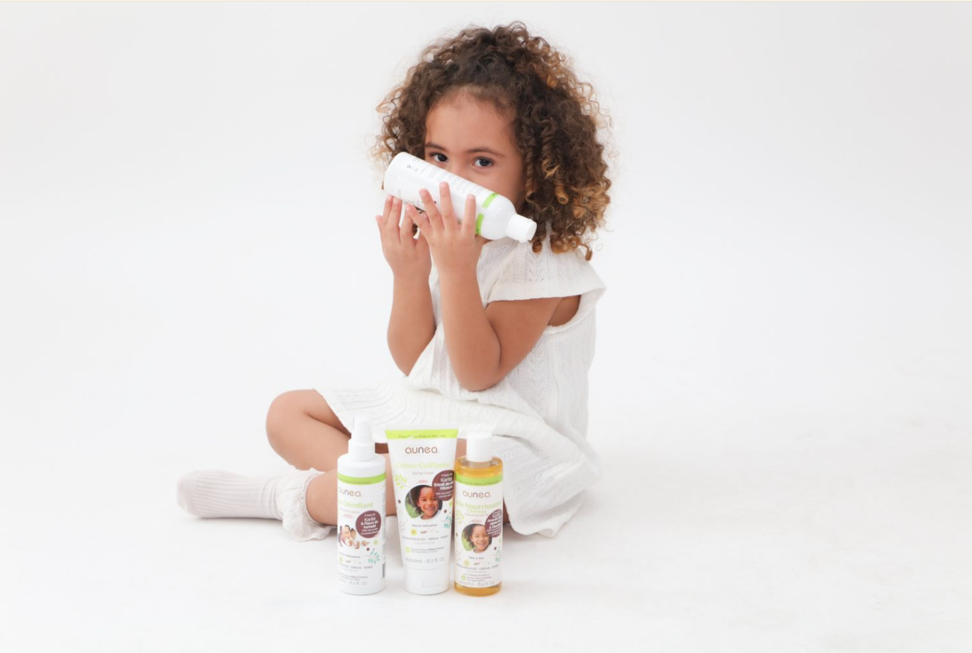 Fille aux cheveux bouclés sentant une bouteille de produit, assise devant d'autres bouteilles sur fond blanc.