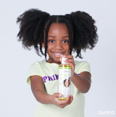 Une jeune fille souriante aux cheveux naturels tient un flacon d'huile capillaire. Elle porte un t-shirt jaune et sourit.