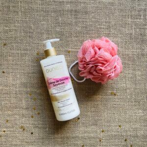 Flacon de gel douche blanc et luffa rose sur une surface beige texturée.