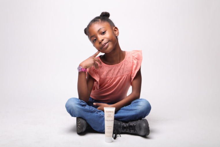 Fille avec les mains sur le visage, assise les jambes croisées à côté d'un tube de produit sur fond blanc.