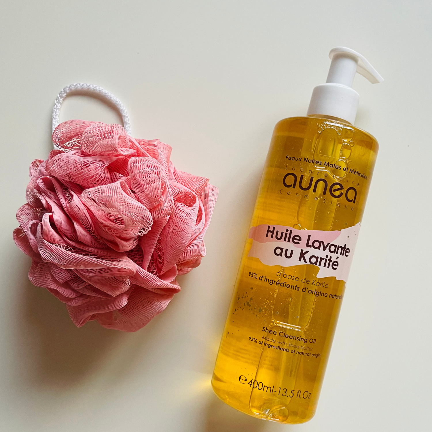 Flacon de gel douche blanc et luffa rose sur une surface beige texturée.