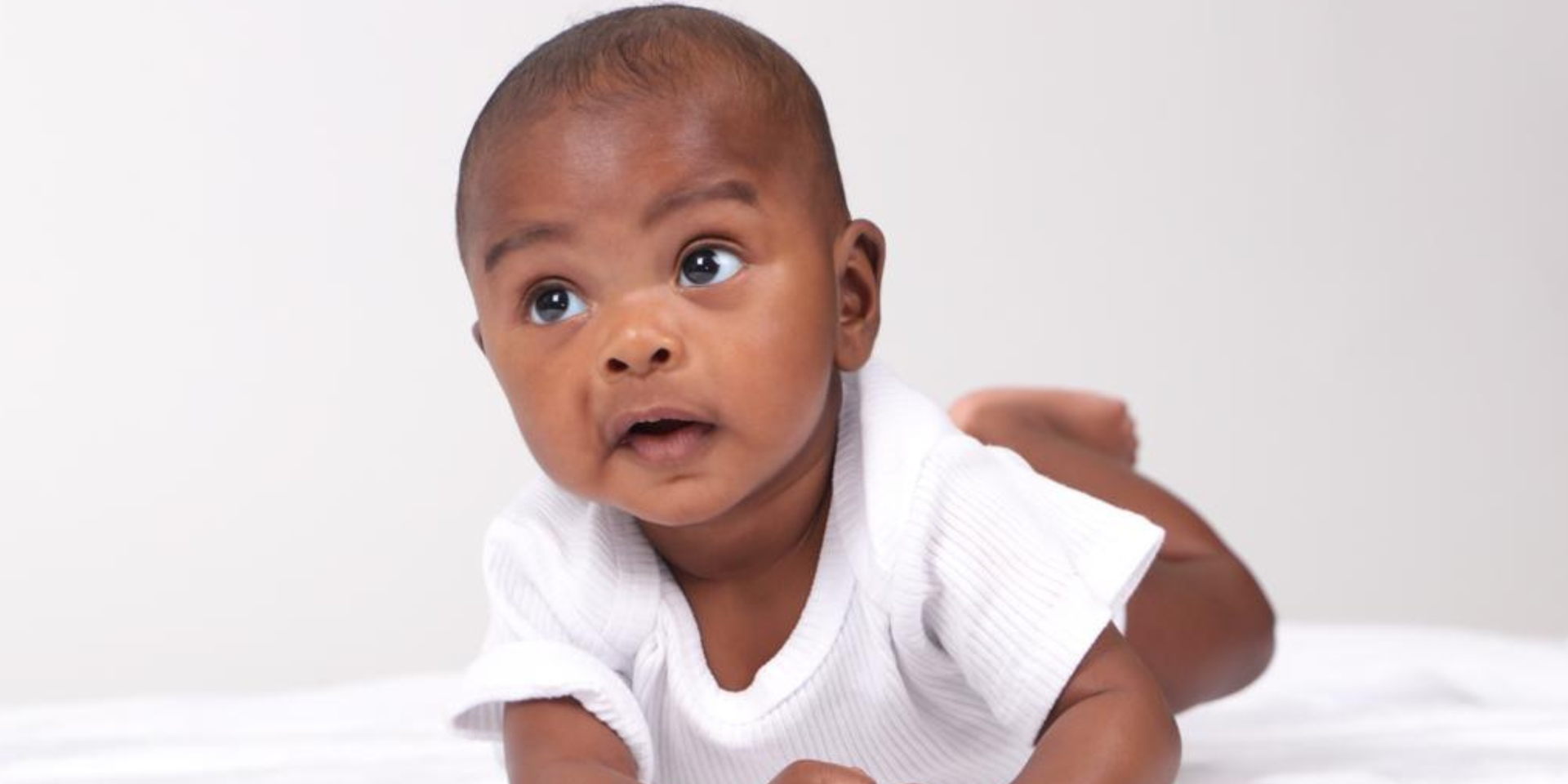 Bébé allongé sur le ventre, regardant vers le haut. Il porte un body blanc.