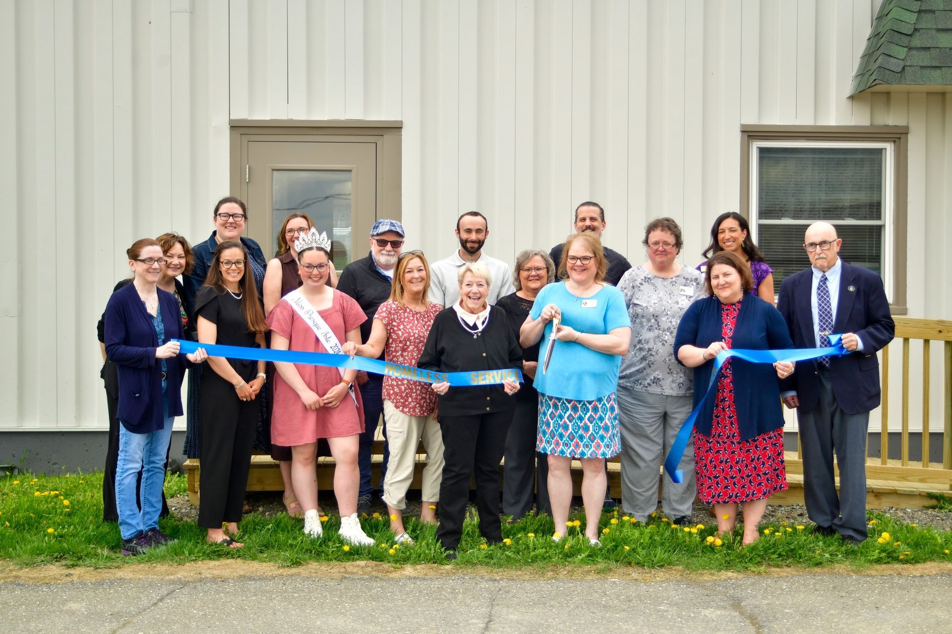 A diverse group of people participating in a ribbon-cutting ceremony in front of a white building.