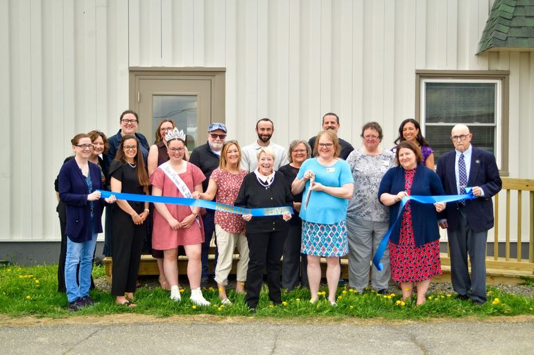 A diverse group of people participating in a ribbon-cutting ceremony in front of a white building.
