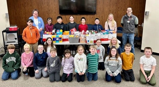 A group of children and adults standing and kneeling behind a table filled with donated food items in a community room.