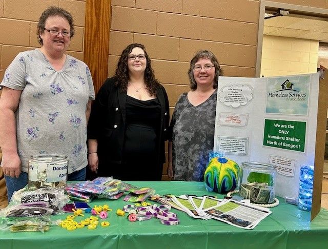 Three people stand behind a table at a booth for Handee Serves, featuring donation jars and informational flyers.