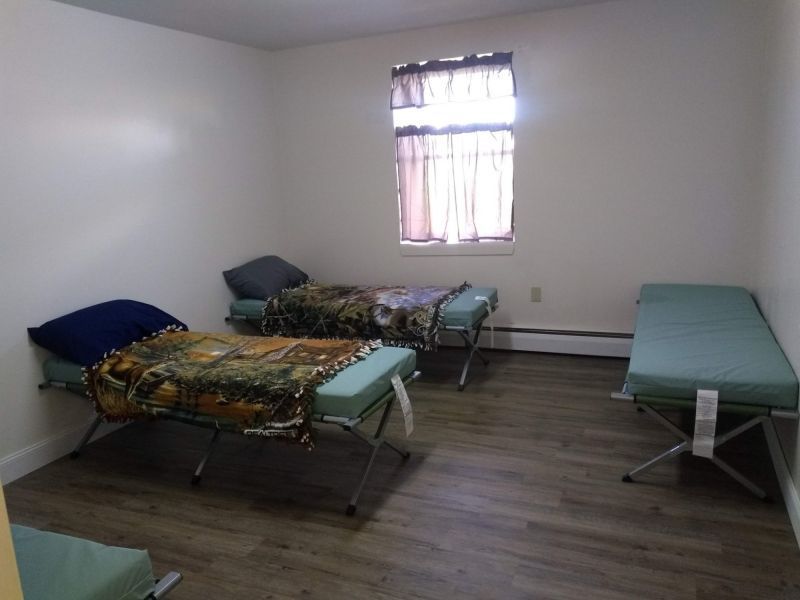 A room with four simple cots, two with blankets and pillows, set on a wood-look floor with a window covered by curtains.