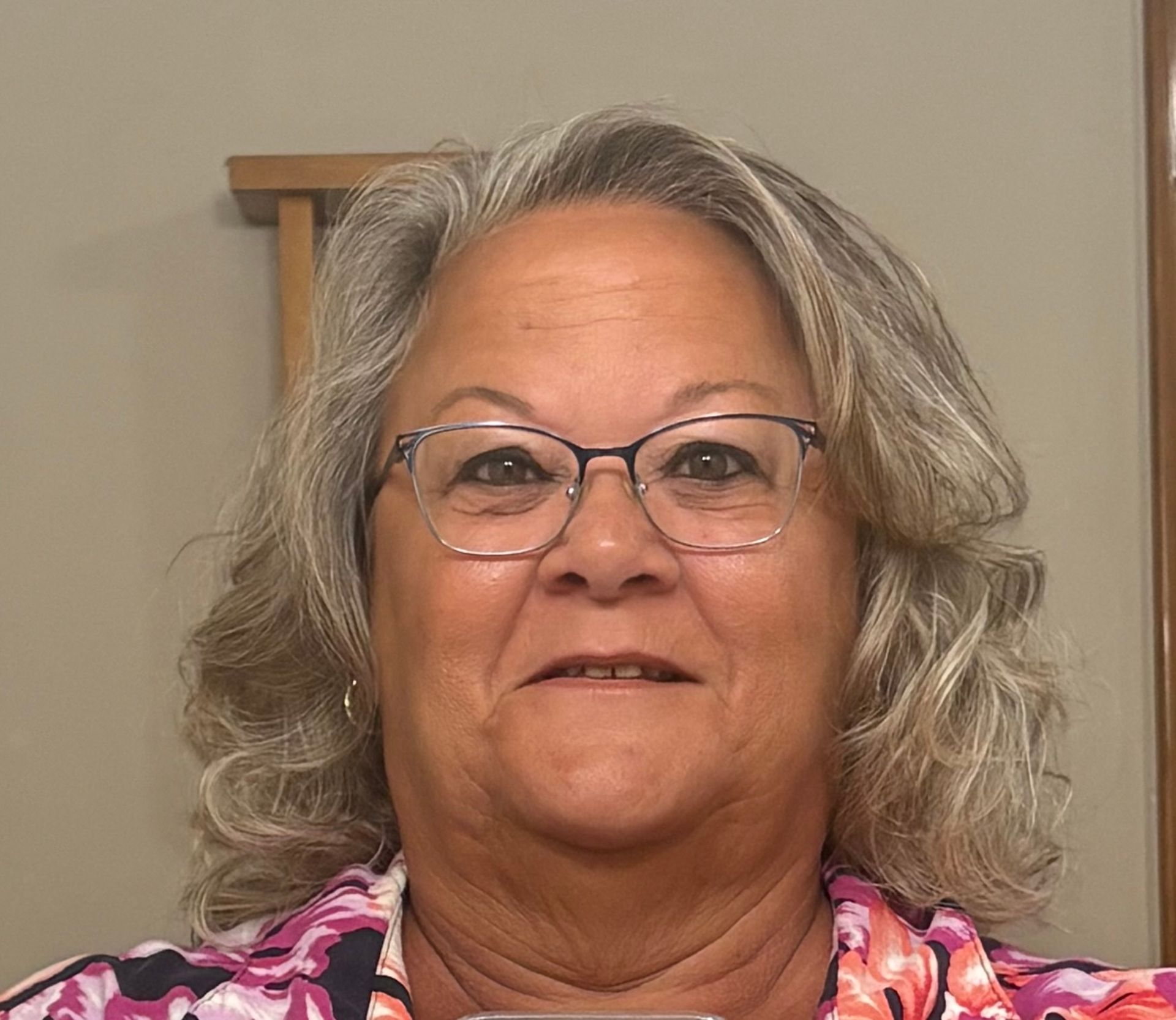 A person with shoulder-length gray hair and glasses, wearing a colorful floral shirt, smiles in a room with neutral walls.