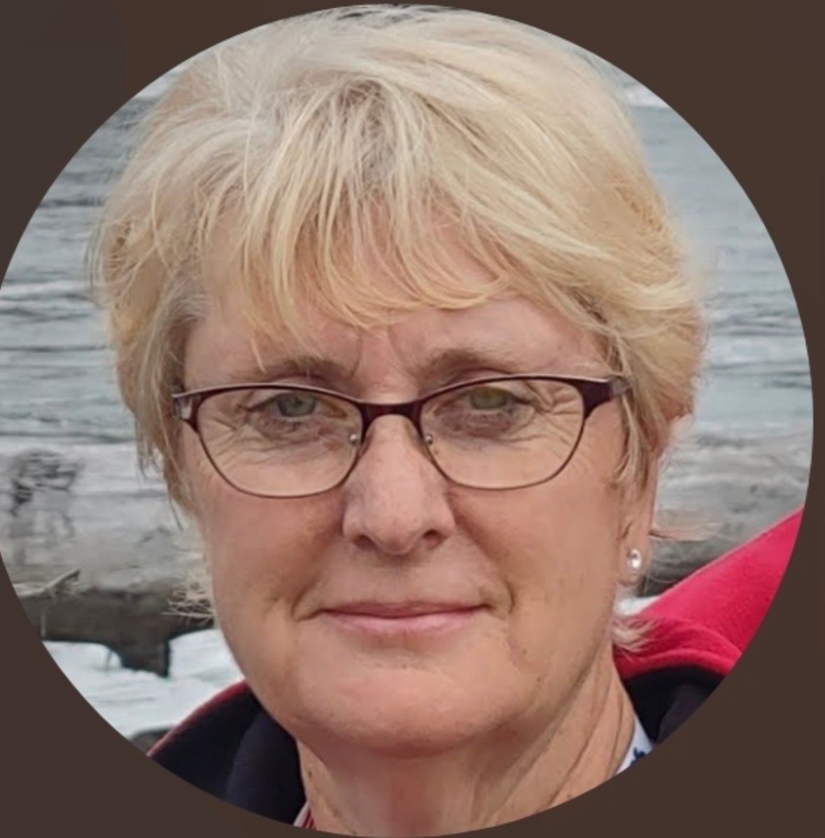 A blonde person wearing glasses, centered in a circular frame against a blurred background of water and snow.