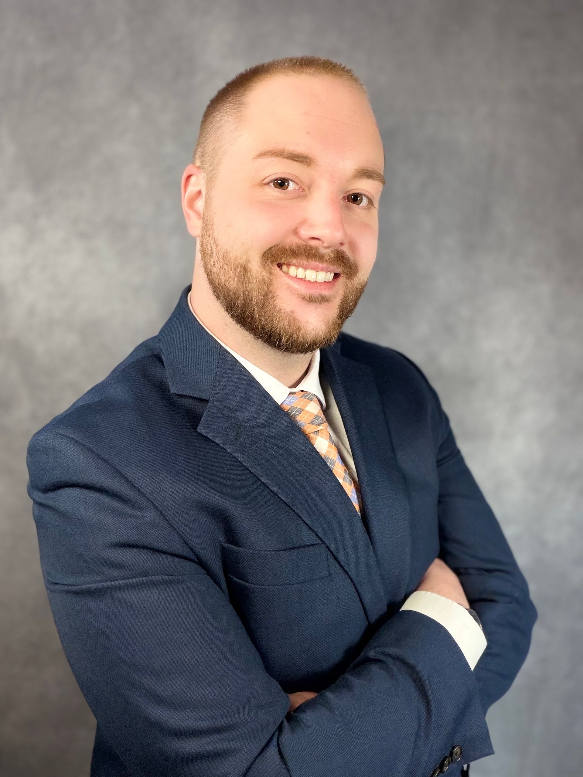 A person in a blue suit jacket, white shirt, and patterned tie, smiling with arms crossed against a grey backdrop.