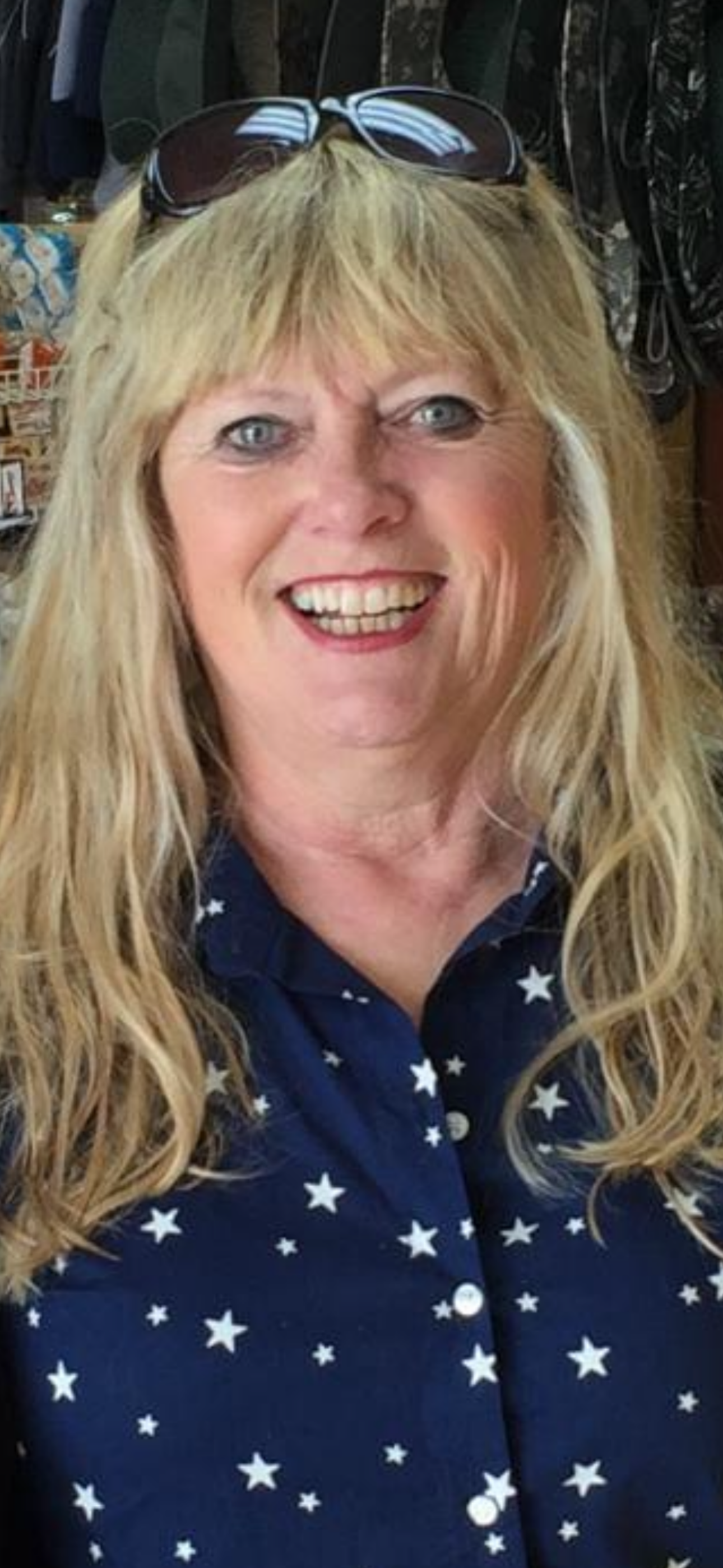 Smiling person with long blonde hair and sunglasses on their head, wearing a blue shirt with white stars.