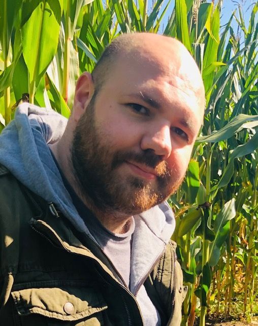 A person with a beard and a hooded jacket stands in front of a field of corn, looking toward the camera.
