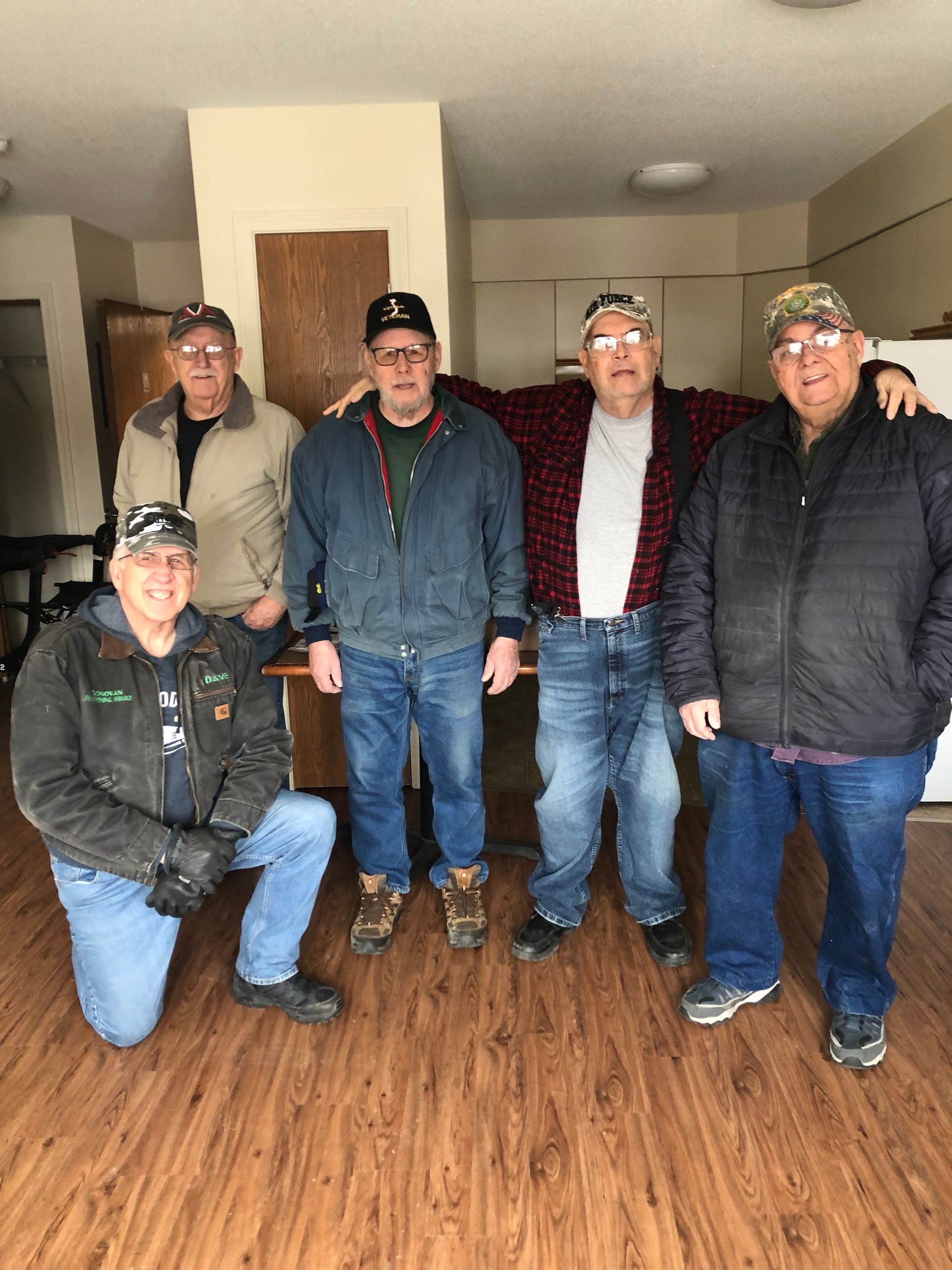 Five people stand and pose in an indoor room with wood-patterned flooring, wearing casual jackets and baseball caps.