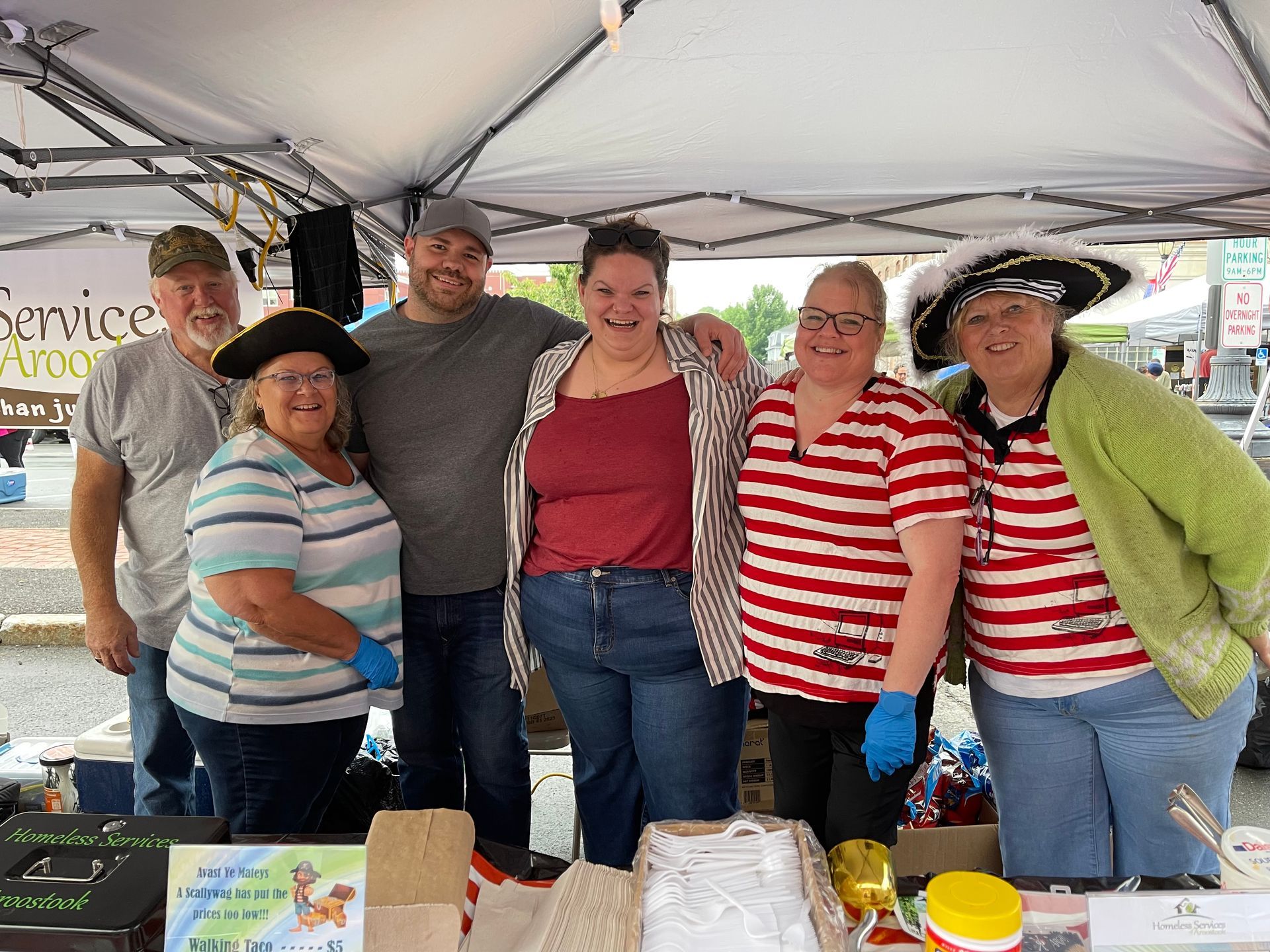 Six people smile under a canopy at a booth, some wearing pirate-themed hats and striped shirts, at an outdoor event.