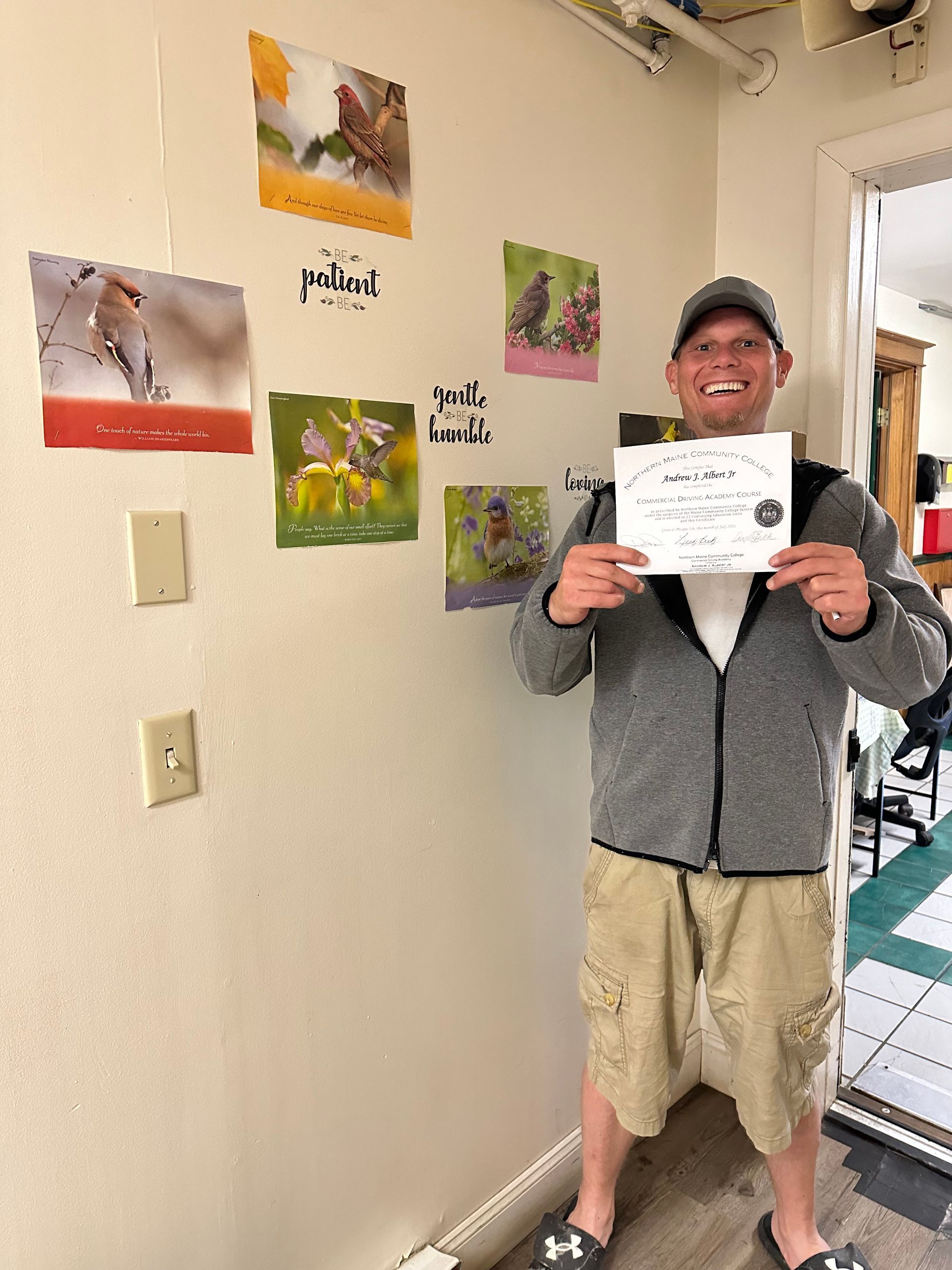 A person smiling and holding a certificate while standing next to a wall decorated with nature photographs.