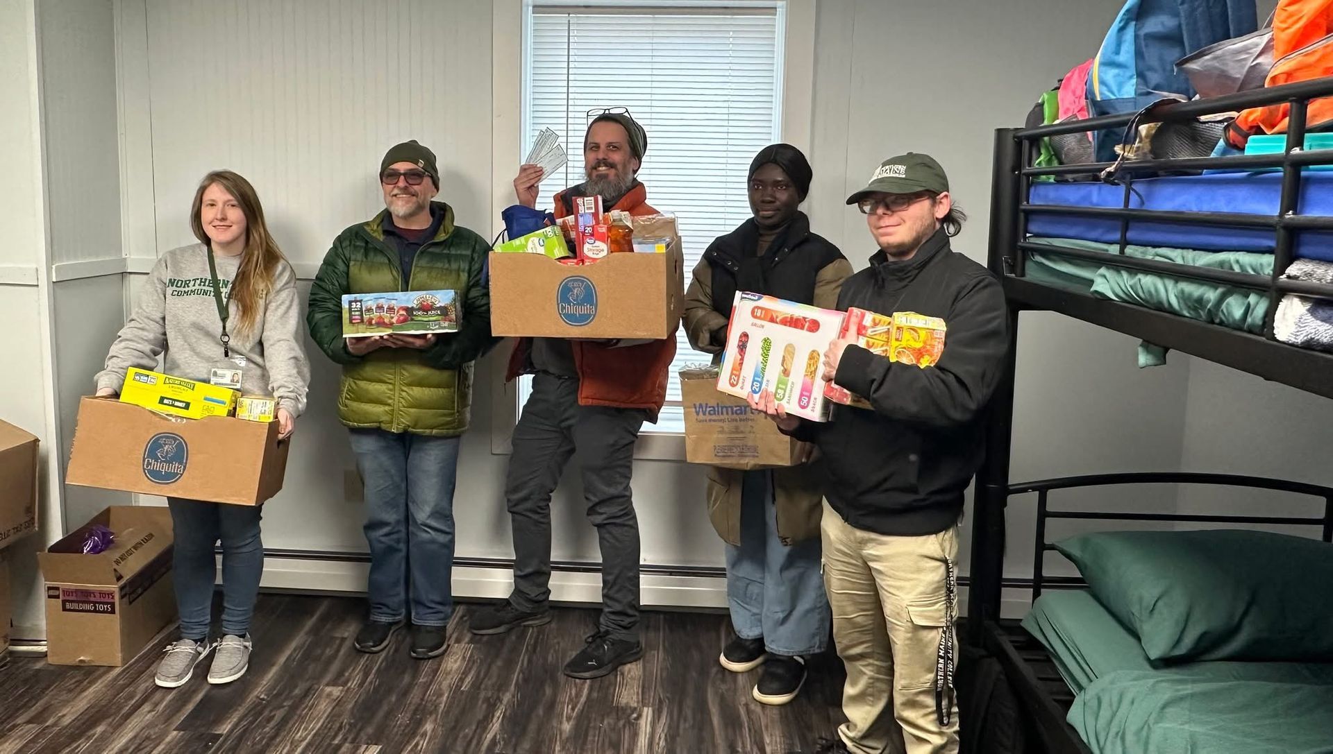 Five people standing in a room with a bunk bed, holding boxes of donated food and supplies for distribution.
