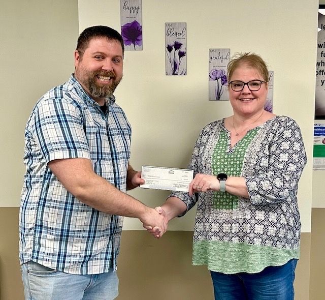 Two people smiling and shaking hands while one hands the other a check in an office setting with floral wall art.