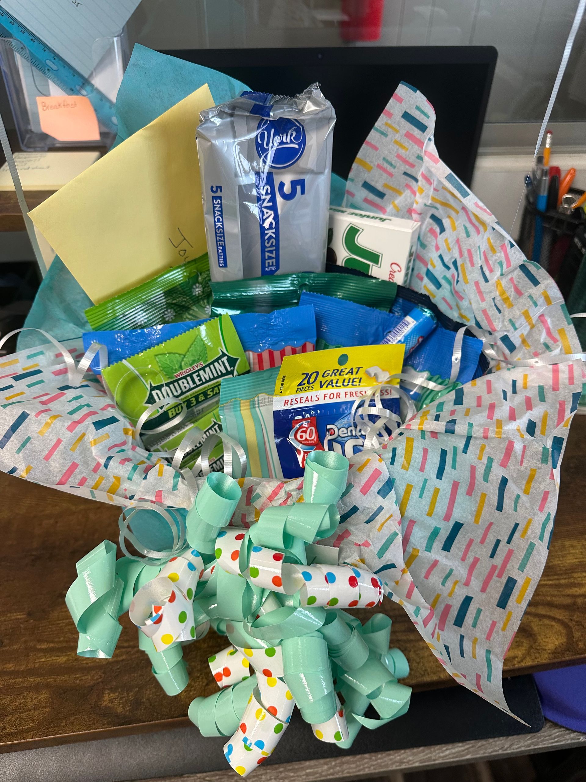 A gift basket filled with snacks and a yellow card, decorated with light green ribbon and colorful confetti tissue paper.