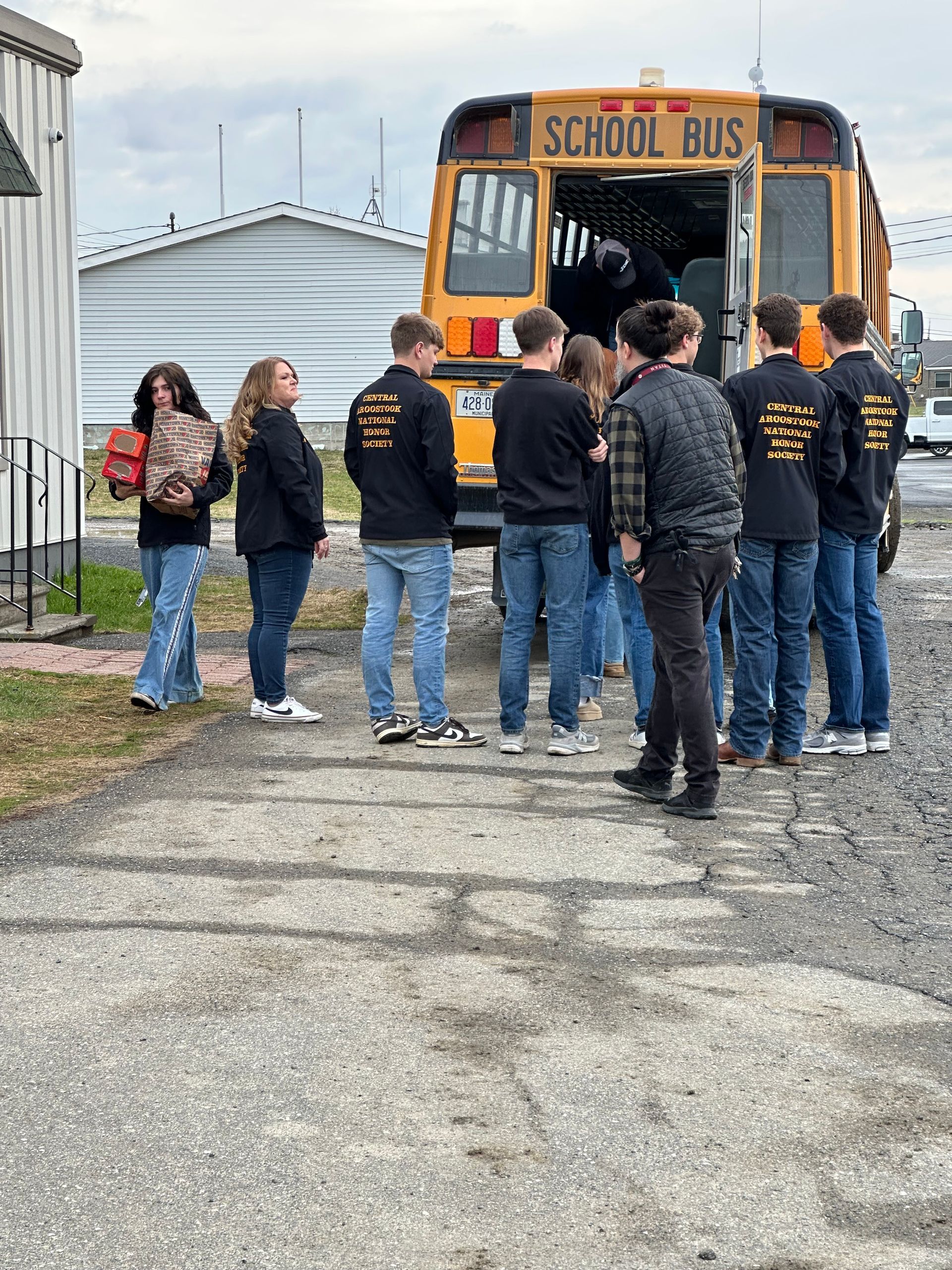 A group of people wearing matching black jackets stand on a gravel surface near the back of an open school bus.